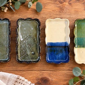 Puede incluir: Cuatro bandejas rectangulares de cerámica con bordes festoneados dispuestas en fila. La primera es de color verde oscuro con textura ondulada, la segunda es verde oliva con superficie texturizada, la tercera es crema y azul, y la cuarta es verde y crema.