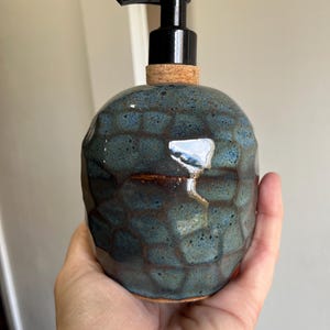 Puede incluir: Dispensador de jabón de cerámica artesanal con un patrón de panal de abeja azul oscuro y marrón. El dispensador tiene una bomba negra y una base de corcho. La botella de cerámica se sostiene en una mano. Altura aproximada: 15 cm.