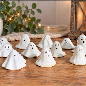 May include: A collection of eleven small, white, glossy ceramic ghost figurines. Each ghost features a smooth, draped shape with two dark, circular eyes and a small, open, circular mouth. The figures are arranged in a group on a wooden surface.