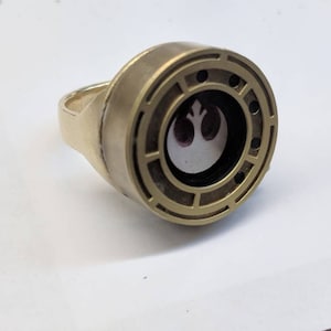 May include: A gold-colored ring with a circular design featuring a white Rebel Alliance symbol inside a clear window. The ring has a textured surface and a raised edge.