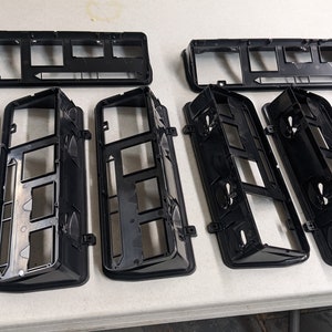 May include: Eight black plastic frames with rectangular cutouts. The frames are arranged in two rows, with four frames in each row. The frames are all the same size and shape.