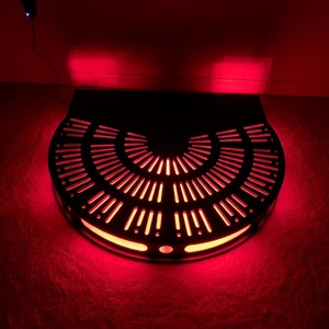 May include: A black metal circular platform with a red light emanating from the edges. The platform has a design of concentric circles with small holes.