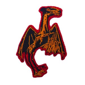 May include: Embroidered patch featuring a stylized dragon design. The patch has a black dragon with orange accents and a red border. The dragon has outstretched wings and a long neck, suitable for jackets, bags, or other fabric items.