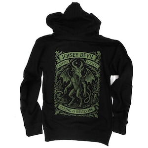 May include: Black hooded sweatshirt featuring a green graphic of the Jersey Devil with text that reads "Jersey Devil Cryptid Society" and "Seeing is Believing". The design includes a horned creature with wings and a tail.