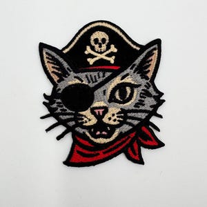 May include: Embroidered patch featuring a gray cat with a black eye patch, wearing a pirate hat with a skull and crossbones. The cat has a red bandana and a smiling expression. The patch is designed with black, gray, red, and tan thread.