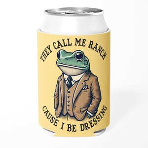 May include: A yellow can cooler with the text "THEY CALL ME RANCH CAUSE I BE DRESSING." The cooler features a cartoon frog wearing a suit and tie. The frog is green with large black eyes and a white collar.