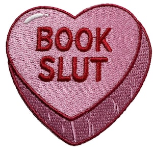 May include: A pink heart-shaped embroidered patch with the words "BOOK SLUT" in red. The patch has a candy heart design with a white accent at the top and a darker pink border.