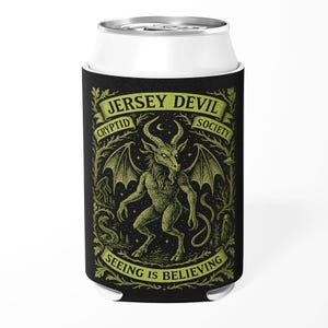 Puede incluir: Enfriador de latas negro con una ilustración verde del Jersey Devil. El texto dice "Jersey Devil Cryptid Society" y "Seeing is Believing". El diseño presenta una criatura cornuda y alada con cola.