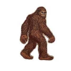 Puede incluir: Parche bordado que representa un Bigfoot marrón caminando. El parche tiene costuras detalladas, mostrando el pelaje y los rasgos faciales de la criatura. El diseño está sobre un fondo blanco, resaltando la forma y los detalles del parche.