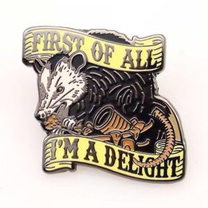 Pode incluir: Um alfinete de metal com um gambá e o texto "FIRST OF ALL I'M A DELIGHT" em faixas amarelas. O gambá é preto, branco e cinza, com um objeto marrom na boca. O alfinete tem uma borda preta.
