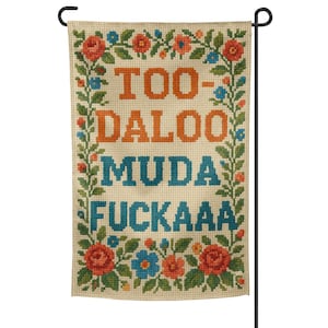 May include: A rectangular garden flag featuring the text "TOO-DALOO MUDA FUCKA" in orange and blue cross-stitch lettering. The flag is decorated with a floral border of red, blue, and orange flowers with green leaves.