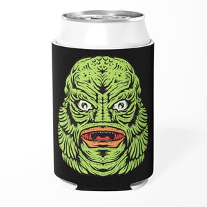 May include: Black can cooler featuring a green monster face with orange lips and white eyes. The monster has a textured, scaly appearance. The can cooler is designed to keep beverages cold.