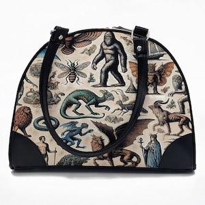 Puede incluir: Un bolso de mano color crema con varias criaturas e ilustraciones míticas. El bolso tiene asas y ribetes negros. Las ilustraciones incluyen un bigfoot, un dragón, un león y una abeja.
