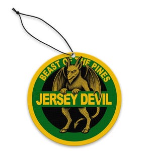 Puede incluir: Ambientador redondo con borde amarillo y verde, con un gráfico amarillo del Jersey Devil. Se muestra el texto "BEAST OF THE PINES" y "JERSEY DEVIL". Una cuerda negra está unida para colgar.