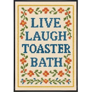 May include: Framed cross-stitch artwork with the words "LIVE LAUGH TOASTER BATH" in blue block letters. The artwork is surrounded by a floral border with orange and green accents, set against a cream background.