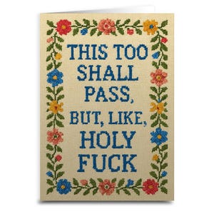 May include: A greeting card with a cream-colored background and blue text that reads "THIS TOO SHALL PASS, BUT, LIKE, HOLY FUCK." The text is in a cross-stitch style, framed by a border of colorful embroidered flowers and green leaves.