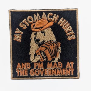 May include: Embroidered patch with a black background and a tan border. The patch features a bear wearing a cowboy hat and playing an accordion. Text reads "MY STOMACH HURTS AND I'M MAD AT THE GOVERNMENT."