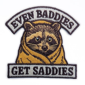 Op de afbeelding: Geborduurde patch met een wasbeer in een gele deken. De patch heeft de tekst "EVEN BADDIES" boven de wasbeer en "GET SADDIES" eronder. De patch is omrand met een donkerbruine kleur.