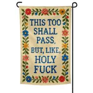 Może przedstawiać: Kremowa flaga ogrodowa z napisem "THIS TOO SHALL PASS, BUT, LIKE, HOLY FUCK" w niebieskim ściegu krzyżykowym. Ma kwiatową obwódkę z czerwonymi, różowymi, żółtymi i niebieskimi kwiatami oraz zielonymi liśćmi.