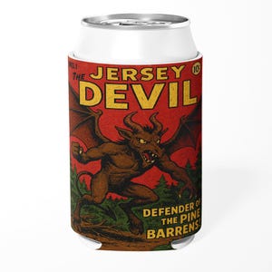 May include: A white can cooler featuring a vintage comic book-style illustration of the Jersey Devil. The artwork depicts a brown, horned creature with bat-like wings against a red background. Text includes "The Jersey Devil" and "Defender of the Pine Barrens."
