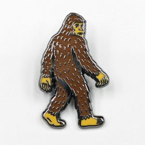 Puede incluir: Un pin de esmalte de Bigfoot marrón con manos y pies amarillos y un contorno plateado. El pin representa a la criatura legendaria caminando hacia adelante, con una textura de pelaje detallada. Un accesorio divertido para mochilas, chaquetas o sombreros.