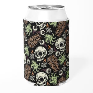 Può includere: Una lattina bianca con un portabevande nero a tema Halloween. Il portabevande presenta un motivo ripetuto di teschi, bare, bulbi oculari, candele e mani di zombi in verde, marrone e bianco. Il design è su sfondo nero.