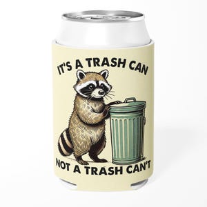 May include: A white can cooler featuring a cartoon raccoon standing next to a green trash can. The text "IT'S A TRASH CAN NOT A TRASH CAN'T" is printed on the cooler. The raccoon is brown and black.
