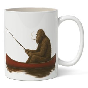 Puede incluir: Taza de cerámica blanca con una ilustración humorística. Representa un Bigfoot marrón fumando un cigarrillo mientras pesca desde una canoa roja. La taza tiene un asa blanca y un interior blanco.