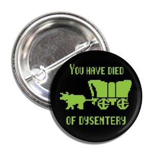 May include: A round, black button with the text "YOU HAVE DIED OF DYSENTERY" in green. The button features a pixelated image of a covered wagon and oxen in green. The button has a silver pin back.