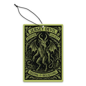 Puede incluir: Un ambientador rectangular con un diseño negro y verde. La imagen presenta una ilustración detallada del Jersey Devil, con el texto "Jersey Devil Cryptid Society" y "Seeing is Believing". Una cuerda negra está adjunta para colgar.