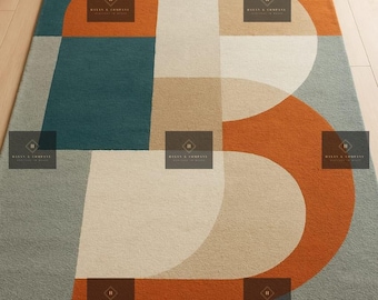May include: A rectangular area rug with a large, stylized letter "B" design. The rug features a color block pattern with teal, orange, beige, and gray sections. The rug is made of a soft, textured material.