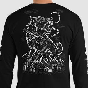Op de afbeelding: Zwart shirt met lange mouwen met een witte illustratie van een grommende wolf die aan een berg is geketend, met een halve maan op de achtergrond. De mouwen hebben witte tekst en kettingontwerpen. Het shirt is gemaakt van zacht materiaal.