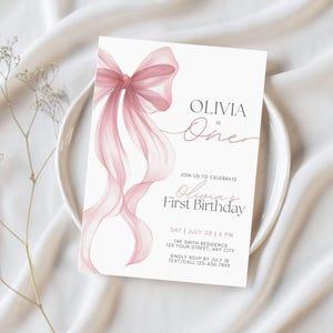 Peut inclure: Une invitation d'anniversaire blanche avec un n&oelig;ud et des rubans roses &agrave; l'aquarelle. Le texte indique "Olivia a un an" et "Premier anniversaire d'Olivia". La carte comprend &eacute;galement la date, l'heure et le lieu de l'&eacute;v&eacute;nement.