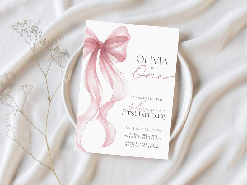 Peut inclure: Une invitation d'anniversaire blanche avec un n&oelig;ud et des rubans roses &agrave; l'aquarelle. Le texte indique "Olivia a un an" et "Premier anniversaire d'Olivia". La carte comprend &eacute;galement la date, l'heure et le lieu de l'&eacute;v&eacute;nement.