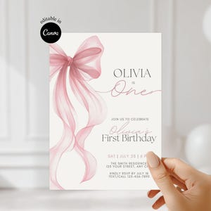 May include: A light-colored invitation card with a watercolor-style pink bow and ribbons. The text reads "OLIVIA is One" and "Olivia's First Birthday". The card includes event details and is held by a hand.