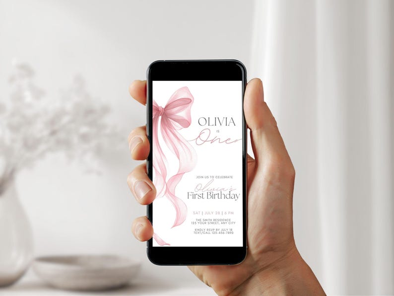 Peut inclure: Un smartphone affiche une invitation rose et blanche pour le premier anniversaire d'Olivia. L'invitation pr&eacute;sente un n&oelig;ud et des rubans &agrave; l'aquarelle. Le texte comprend la date, l'heure et le lieu.