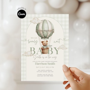 Peut inclure: Invitation de baby shower avec un ballon à air chaud aquarelle, un ours en peluche et le texte "We can bearly wait BABY". L'invitation est dans des tons verts et beiges doux et comprend les détails de l'événement.