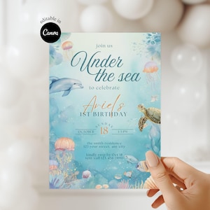 Puede incluir: Una tarjeta de invitación con el tema "Bajo el mar". El diseño presenta un delfín, una tortuga marina, medusas y otras criaturas marinas. El texto dice "Únete a nosotros Bajo el mar para celebrar el 1er cumpleaños de Ariel" con la fecha y la hora.