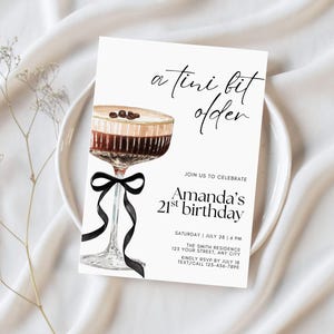 May include: A white invitation card with the text "a tini bit older" and "Amanda's 21st birthday". The card features a watercolor illustration of a cocktail with coffee beans and a black ribbon. The invitation is on a white plate.