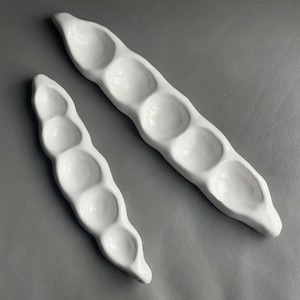 May include: Two white ceramic serving dishes shaped like pea pods. The larger pod has five sections, and the smaller one has three. The dishes are on a grey surface.