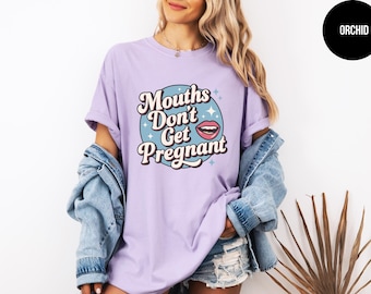 May include: A lilac-colored t-shirt with the text "Mouths Don't Get Pregnant" in a retro font. The design includes a blue circle with a mouth graphic and star accents. The model is wearing a denim jacket.