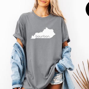 Peut inclure: T-shirt gris avec un contour blanc de l'État du Kentucky et le mot "bourbon" imprimé en dessous. Le t-shirt est porté avec une veste en jean et un short.