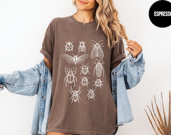 May include: Espresso-colored t-shirt featuring a white insect illustration. The graphic includes various insects such as beetles, moths, and butterflies. The shirt is worn with a denim jacket.
