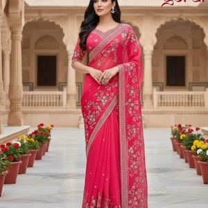 May include: A vibrant pink saree with intricate floral embroidery and a delicate border. The saree features a matching blouse with short sleeves and a V-neckline. The word "goral" is visible in the upper right corner.