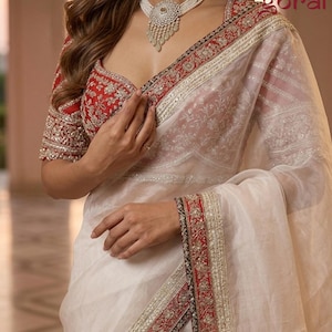May include: A cream saree with a red and gold embroidered border. The saree is worn with a red blouse featuring detailed embroidery on the sleeves and neckline. A pearl necklace with a silver pendant is worn.