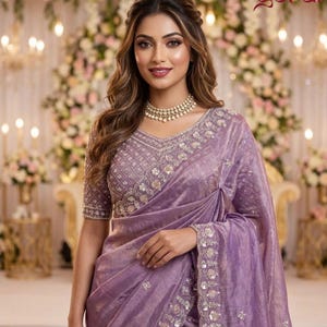 May include: A woman in a lavender sari with detailed embroidery and embellishments. The sari has a floral pattern on the borders and bodice. The background features floral arrangements and soft lighting, suggesting a formal occasion.
