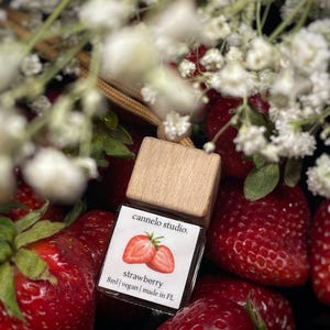 May include: A small glass bottle with a wooden cap, labelled "cannelo studio." The label features a strawberry illustration and the word "strawberry." The bottle is surrounded by fresh, ripe strawberries and white flowers.