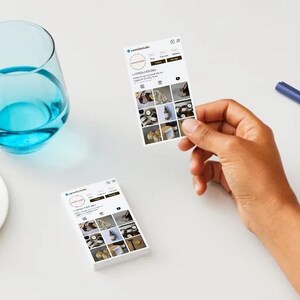 May include: A still-life image featuring a glass of blue liquid, a plate with macarons, and two business cards. One card is held by a hand, displaying a social media profile with product images. A blue pen rests on the white surface.