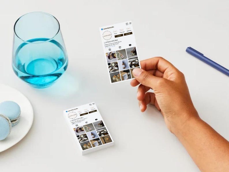 May include: A still-life image featuring a glass of blue liquid, a plate with macarons, and two business cards. One card is held by a hand, displaying a social media profile with product images. A blue pen rests on the white surface.