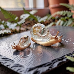 May include: Two snail figurines with smooth, matte gold-toned bodies and clear, rounded shells. The larger shell contains embedded metallic elements, while the smaller shell features gold-colored flecks. Both snails have two distinct eye stalks.